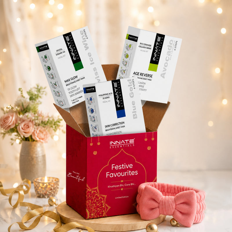 A red Innate Essentials gift box labeled 'Festive Favourites' filled with boxed skincare products, styled with a pink bow headband, gold ribbons, flowers, and warm string lights in the background.